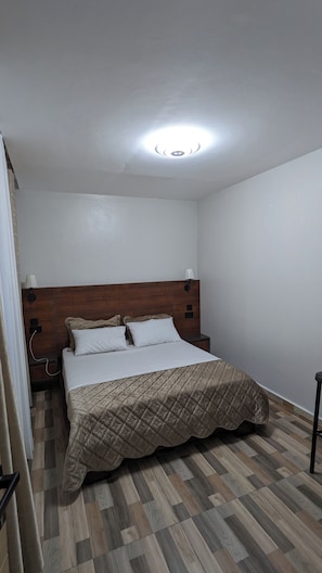 A bedroom with a wooden headboard, a bed with a quilted cover, a nightstand, a wall-mounted lamp, and a ceiling light. - Cozy One bedroom apartment in Kigali , Rebero with panoramic mountain views (Kigali)