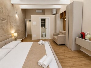 Una habitación de hotel con cama, zona de estar con una silla, una mesita con tetera y un espejo.