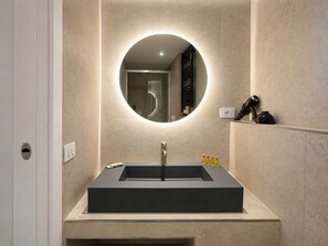 Un baño moderno con un lavamanos rectangular, un espejo redondo y un estante empotrado con artículos de aseo.
