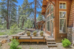 A wooden cabin with a deck, large windows, and a stone wall. - Old Greenwood Cabin 01 (Truckee)