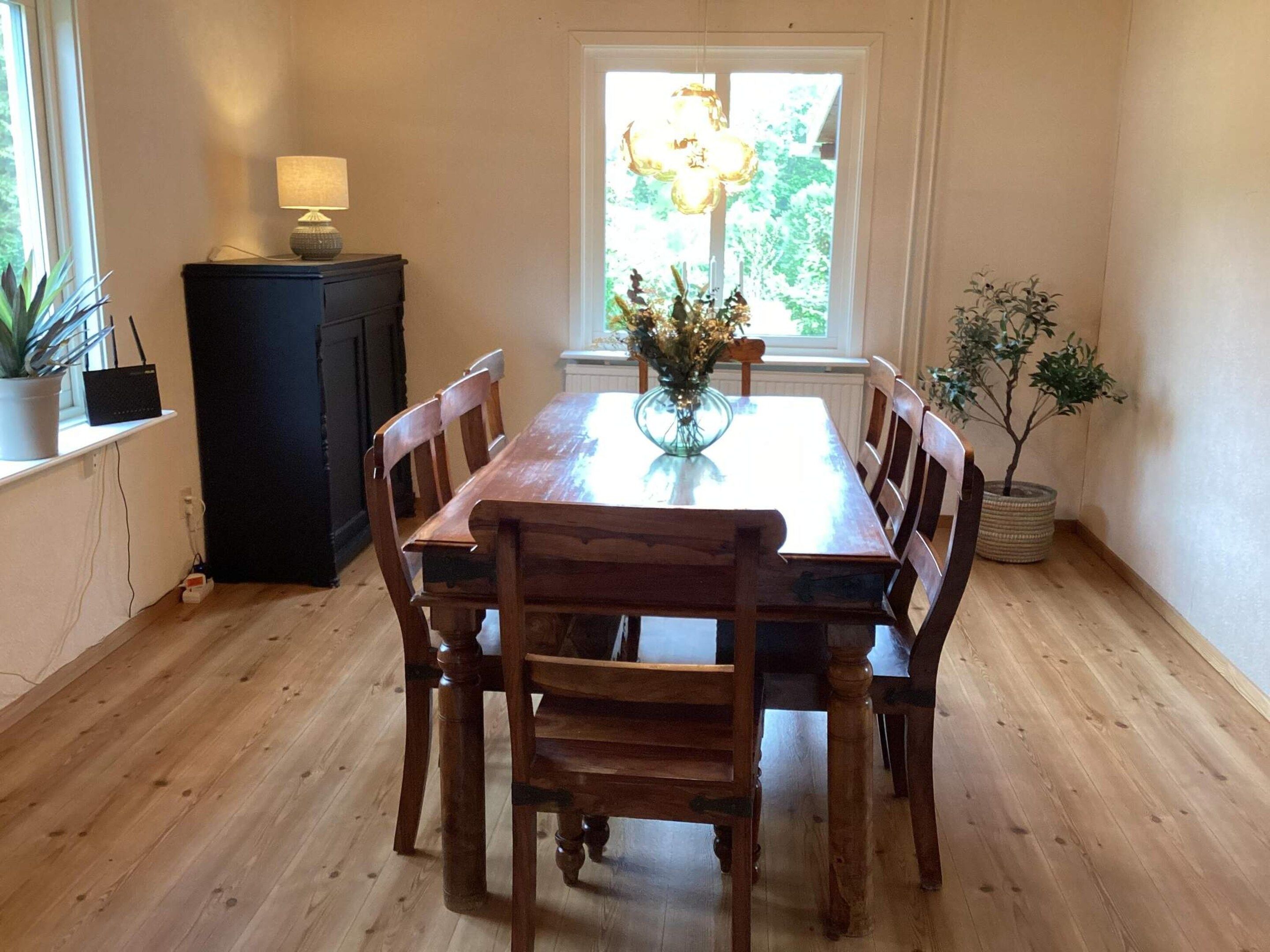 A dining area with a wooden table and chairs, a vase with flowers, and a window with a view of greenery.