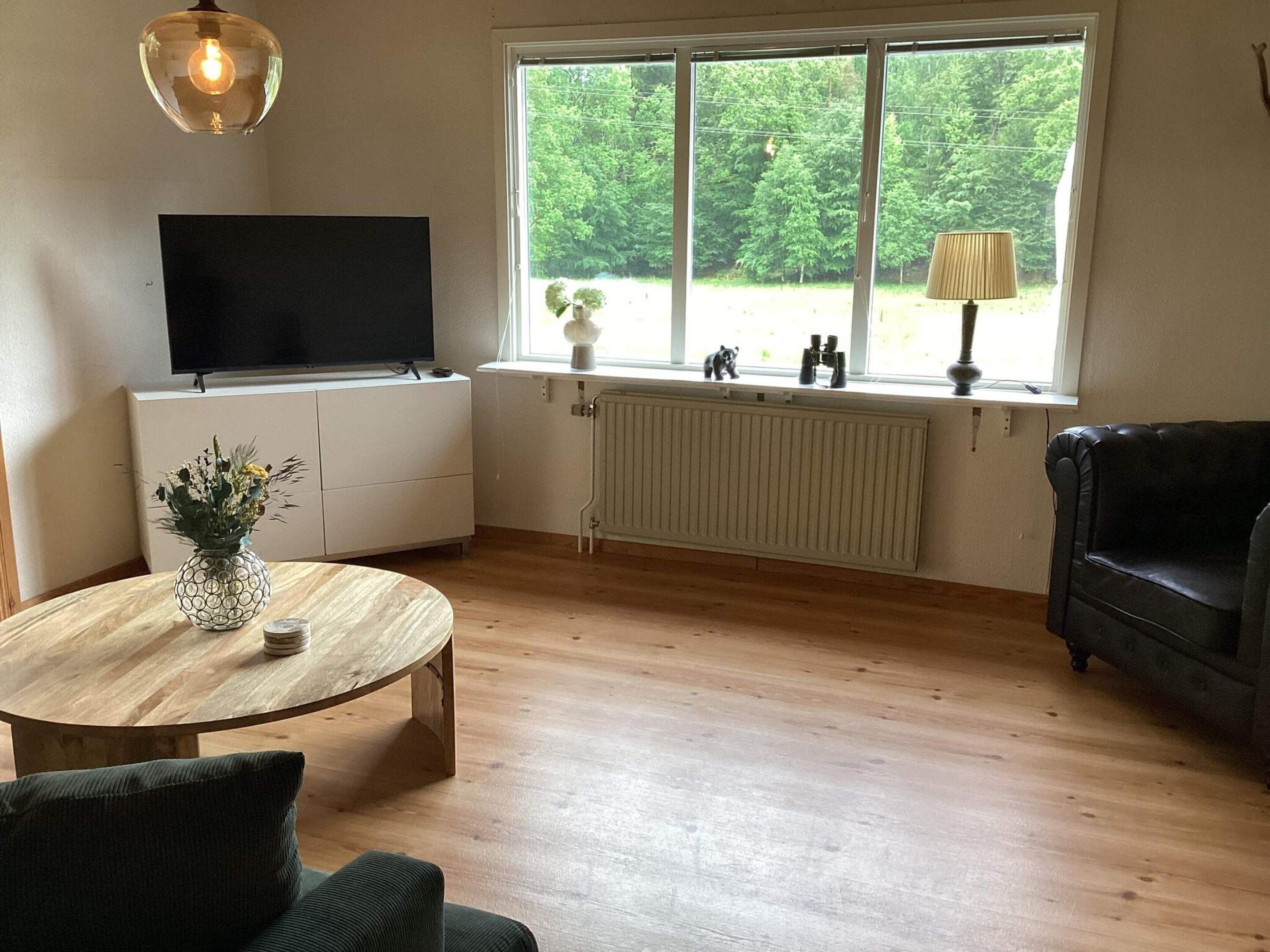 A living room with a wooden floor, a round coffee table, a sofa, a TV on a white cabinet, and a window with a view of greenery.