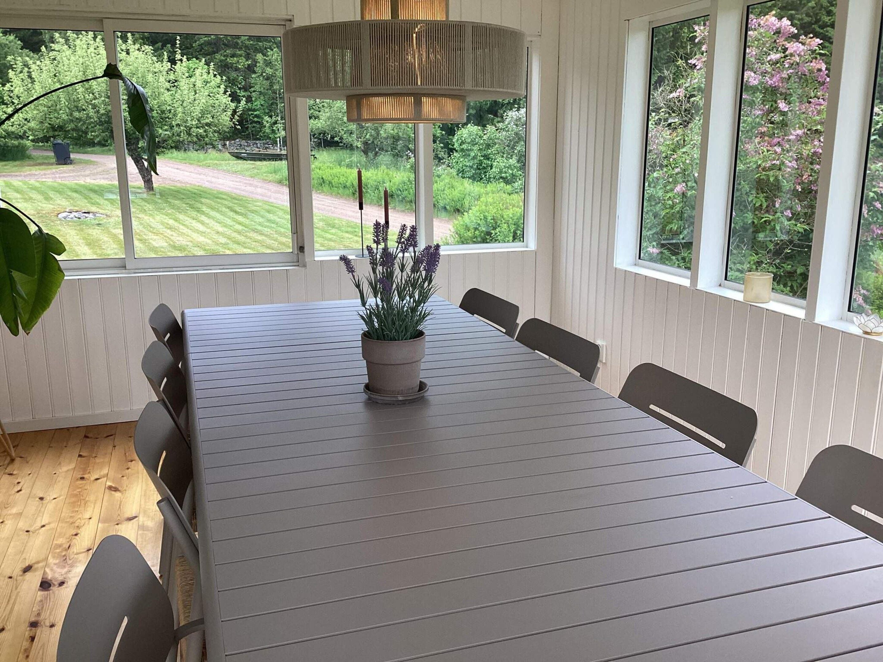 A dining area with a long table, chairs, a potted plant, and large windows with a view of greenery.