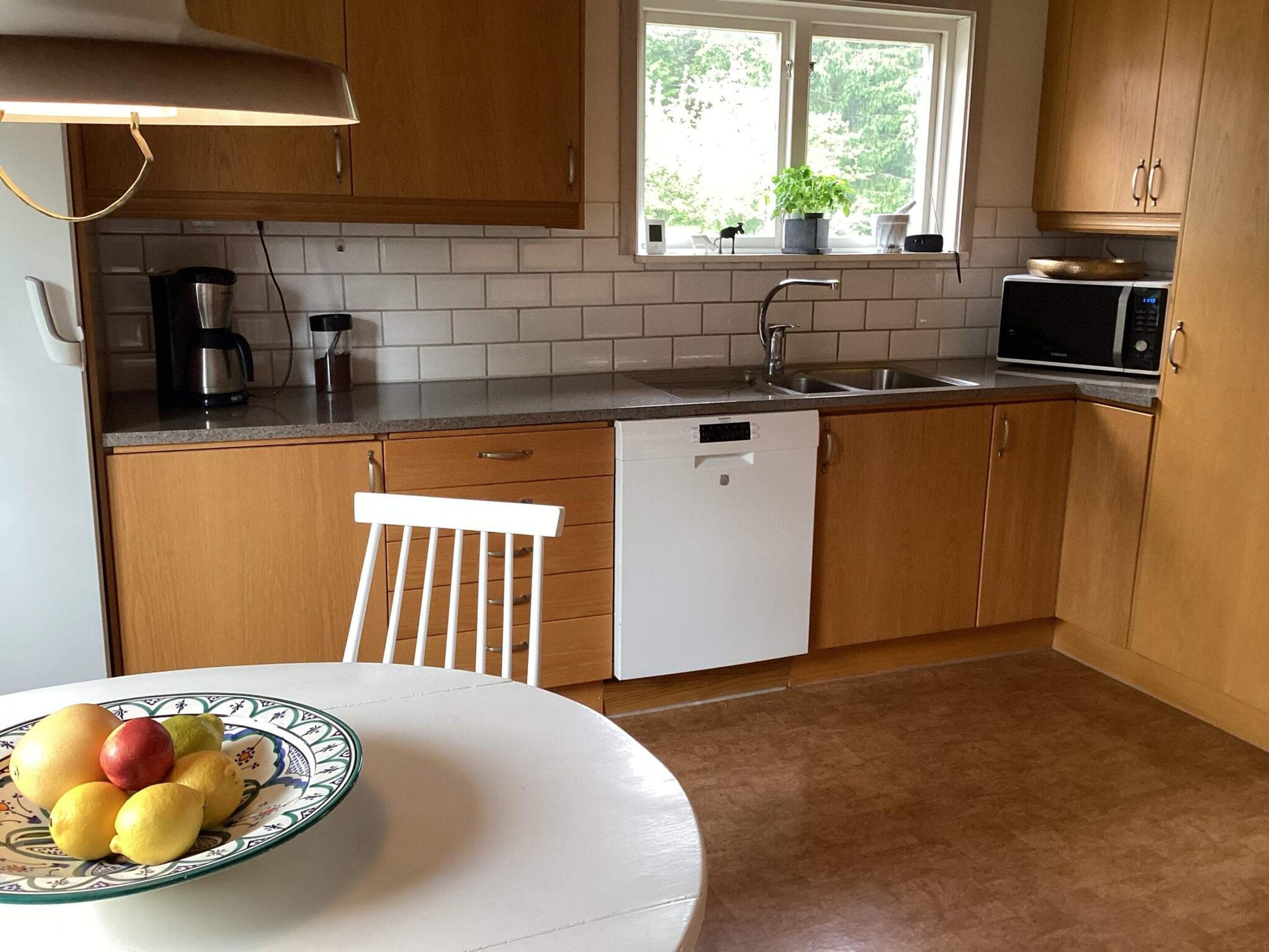 A kitchen with wooden cabinets, a white countertop, a sink, a dishwasher, a microwave, and a window with a plant.
