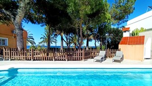 Una piscina cristalina con sillas de descanso, una valla de madera y árboles al fondo.