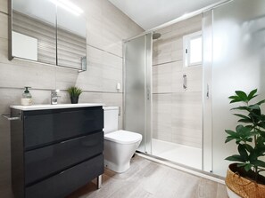 Une salle de bain moderne avec un meuble-lavabo de couleur foncée, des toilettes blanches, une douche à parois de verre et une plante en pot.