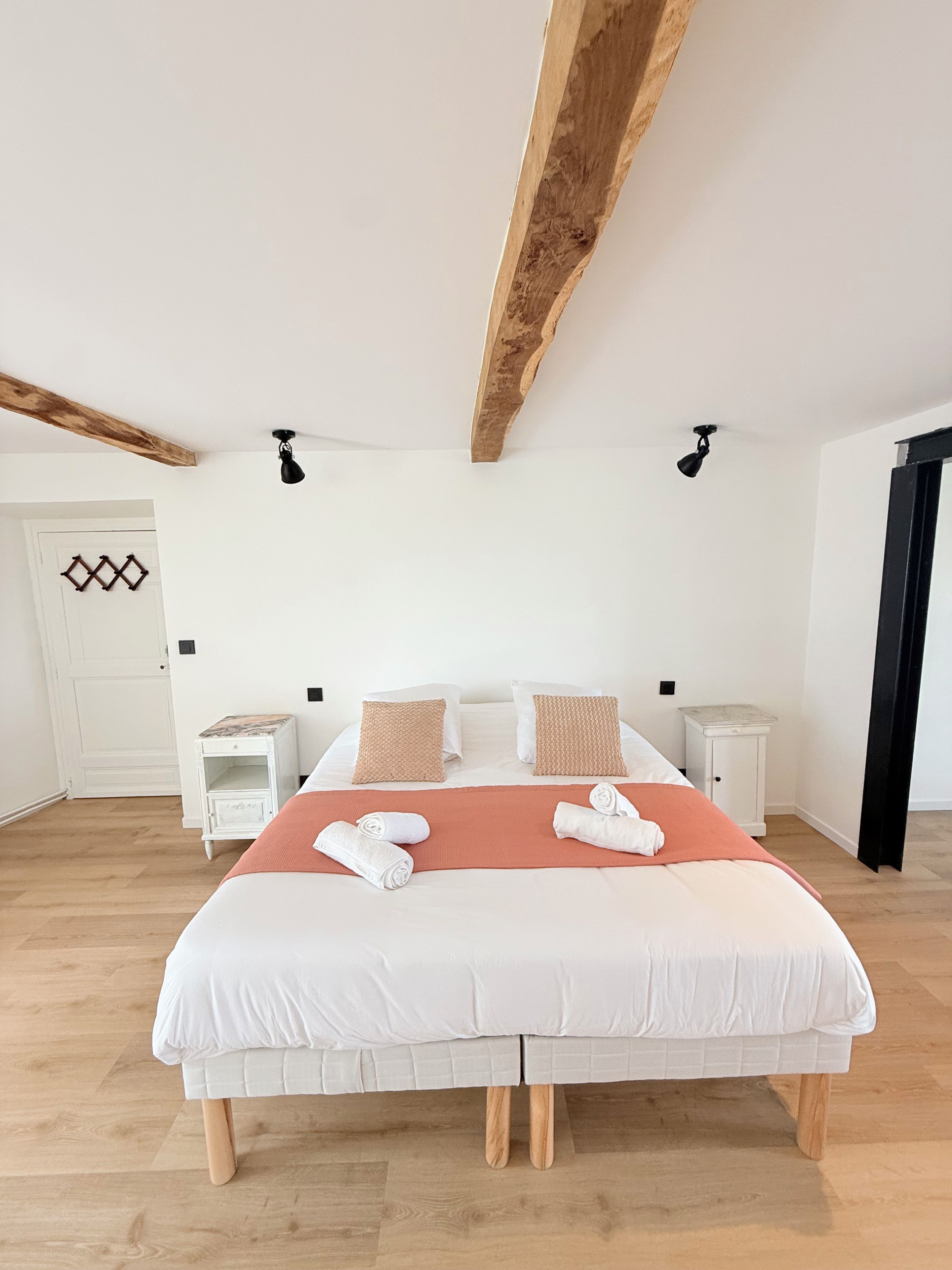 Ein Schlafzimmer mit einem großen Bett, zwei Nachttischen, einem Holzbalken an der Decke und einer weißen Tür mit geometrischem Muster.