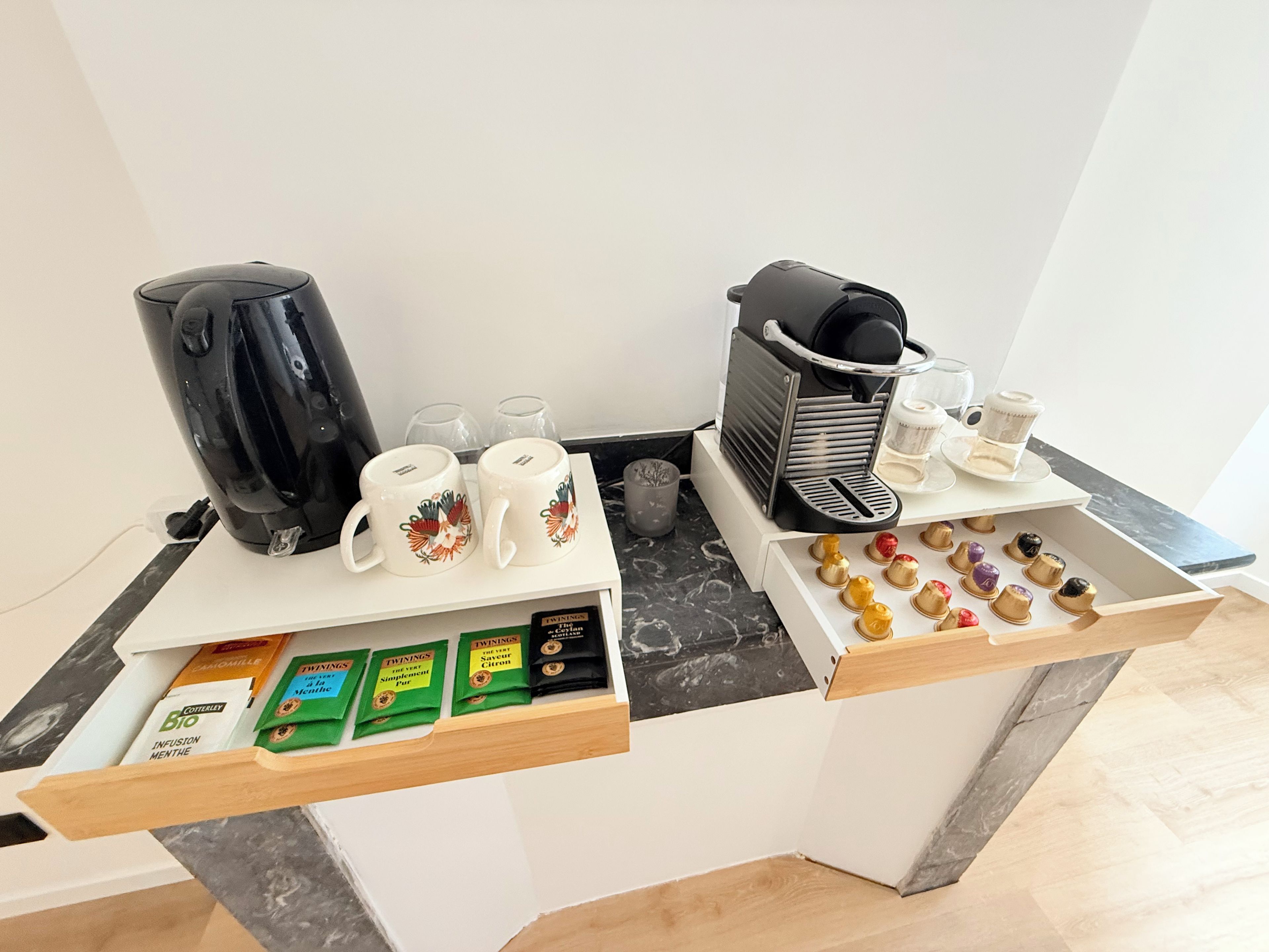 Een koffiehoek met een koffiezetapparaat, twee mokken, een koffiemolen en een lade met koffiepads.