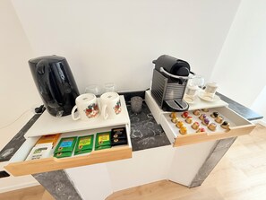 Una estación de café con una cafetera, dos tazas, un molinillo de café y un cajón con cápsulas de café.