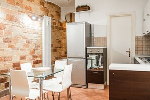 Uma cozinha compacta com parede de tijolos, geladeira, forno embutido e um espaço de refeições com cadeiras e mesa de vidro.