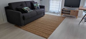 A living room with a black sofa, a brown rug, a TV, and a wooden side table. - Recent 25m2 studio with parking space (Aytré)