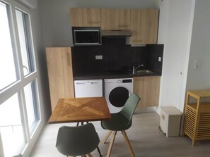 A compact kitchen with a microwave, dishwasher, and washing machine, wooden cabinets, a sink, and a dining area with a table and chairs. - Recent 25m2 studio with parking space (Aytré)