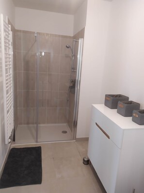 A bathroom with a glass-enclosed shower, a white vanity, and storage baskets. - Recent 25m2 studio with parking space (Aytré)