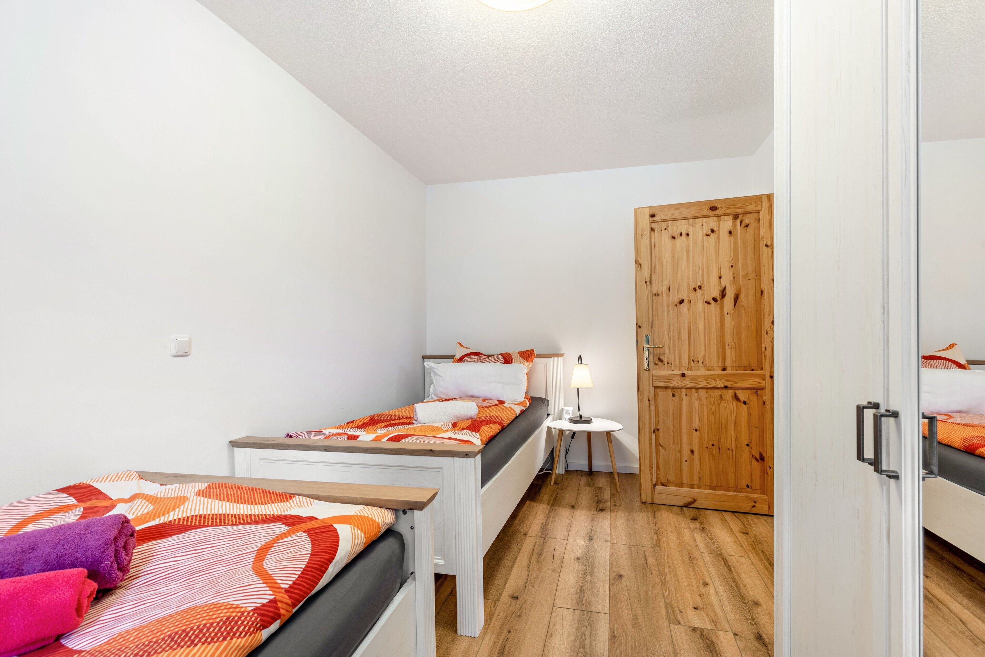 A small, well-lit room with two beds, a wooden wardrobe, and a small table with a lamp.