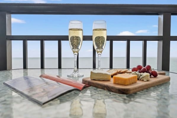 Uma mesa com duas taças de champanhe, um tabuleiro de queijos com diversos tipos e uvas, e uma revista numa varanda com vista para o mar.