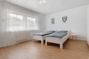 Une chambre avec deux lits, une fenêtre avec des rideaux légers, un radiateur et deux œuvres d’art accrochées au mur.