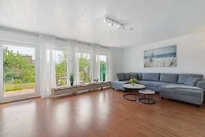 A spacious living room with a grey sofa, a round coffee table, and large windows with white curtains. - Holiday Home 'Urban 2.0' with Terrace and Private Garden (Büttelborn)