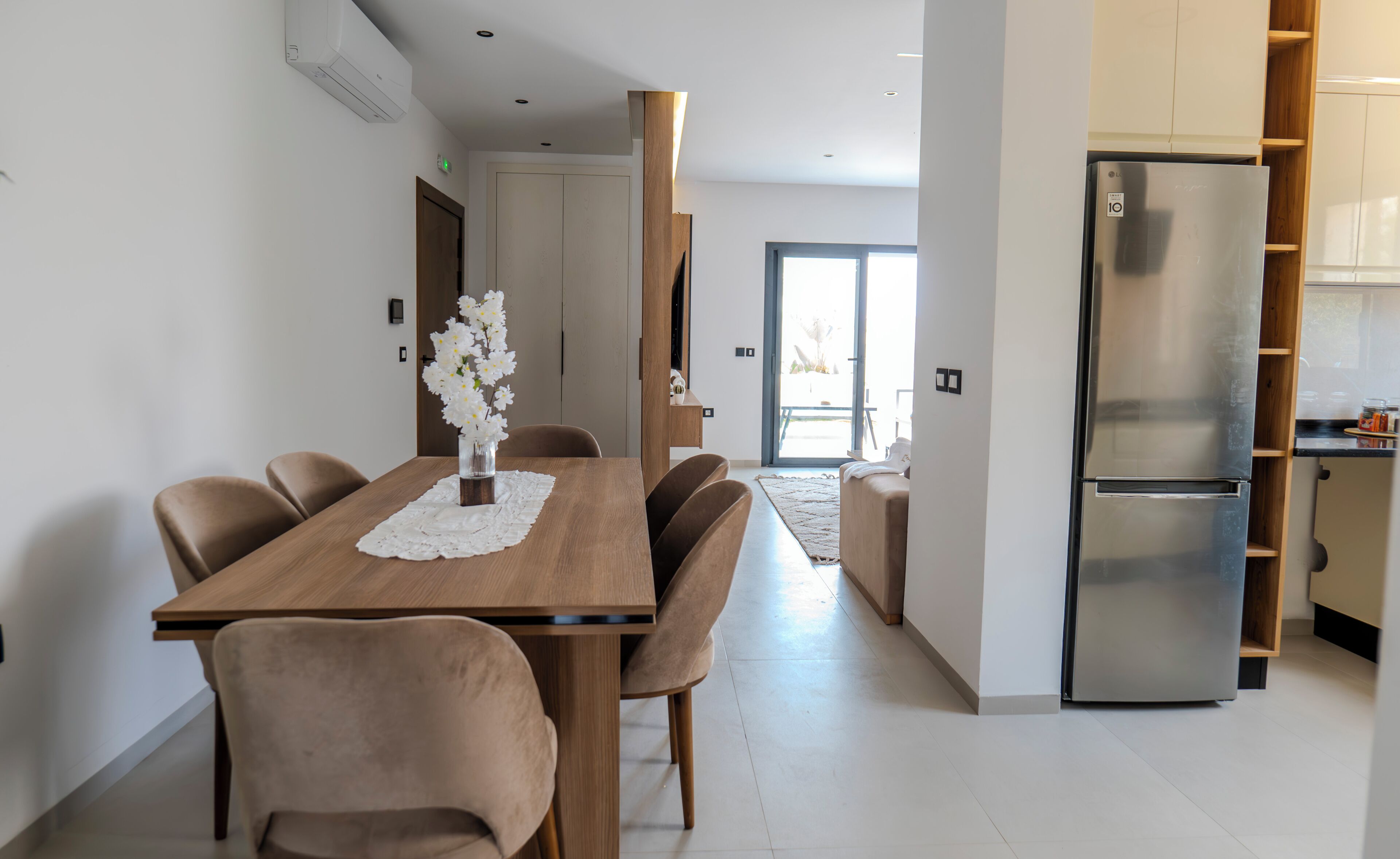 Une cuisine moderne dotée d’un réfrigérateur en acier inoxydable, un coin repas avec une table et des chaises en bois, et un couloir menant à un balcon.