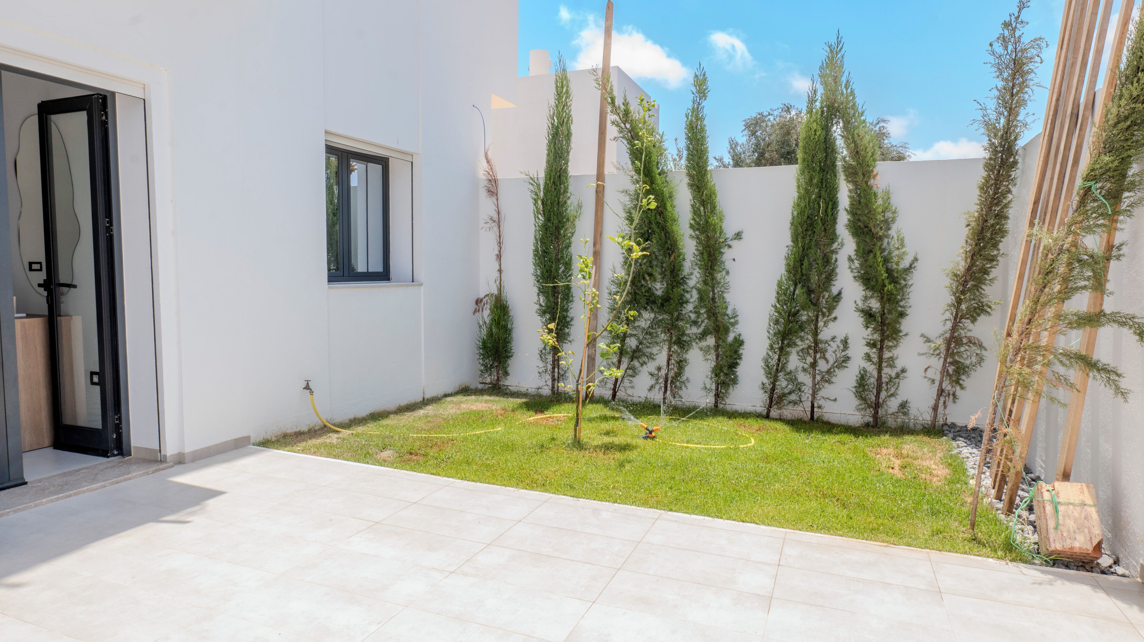 Il s’agit d’un bâtiment moderne à l’extérieur blanc, doté d’une petite fenêtre et d’une porte. On y trouve une zone herbeuse avec de jeunes arbres et un tuyau d’arrosage.