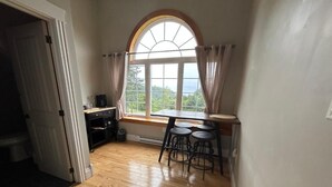 A room with a wooden floor, a window with curtains, a table with chairs, and a door leading to another room. - Tides Inn Guest Suites (Halls Harbour)