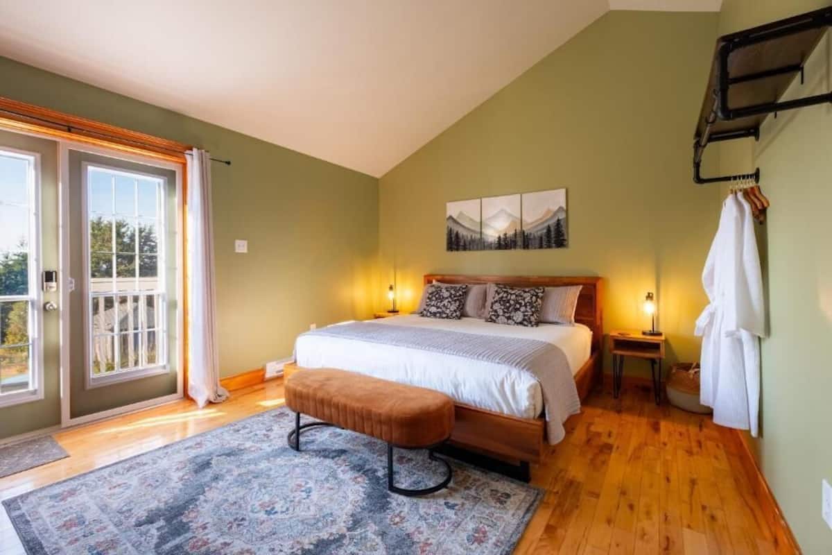 A bedroom with a bed, bench, bedside tables, a painting, and a window with curtains.