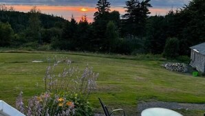 A patio with a round table and chairs, a white railing, and a view of a grassy field and trees. - Tides Inn Guest Suites (Halls Harbour)