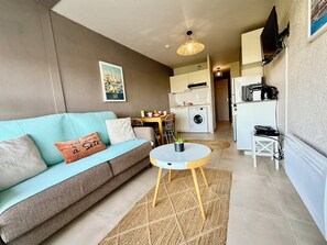 A compact living space with a sofa, a small table, a washing machine, and a kitchenette.