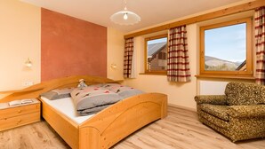 Quarto com cama de madeira, mesa de cabeceira, poltrona estampada, janela com cortinas e vista para um edifício.