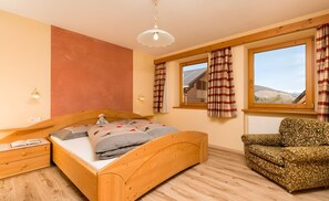 Ein Schlafzimmer mit einem hölzernen Bett, einem Nachttisch, einem gemusterten Sessel, einem Fenster mit Vorhängen und Blick auf ein Gebäude.