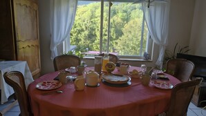 Une table à manger dressée avec différents plats et boissons, entourée de chaises en osier, avec vue sur les arbres à travers une fenêtre.
