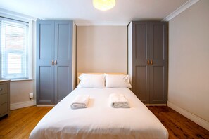 Quarto com cama, dois armários cinza, mesa de cabeceira e janela com persianas.