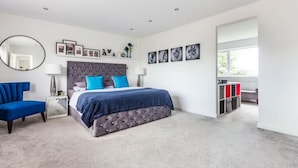 A bedroom with a tufted headboard bed, a blue chair, a bedside table with a lamp, a large mirror, and a window with a view.