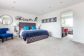 A bedroom with a tufted headboard bed, a blue chair, a bedside table with a lamp, a large mirror, and a window with a view.