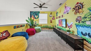 Ein Zimmer mit einer Wand im Super-Mario-Design, einem Deckenventilator, Sitzsäcken, einem Fernseher und einem Fenster mit Jalousien.