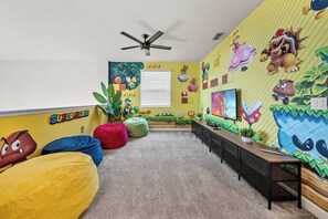 Ein Zimmer mit einer Wand im Super-Mario-Design, einem Deckenventilator, Sitzsäcken, einem Fernseher und einem Fenster mit Jalousien.