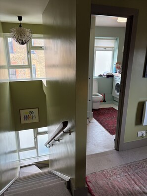 Eine Treppe führt zu einer Empore mit einem gerahmten Bild, einer Pendelleuchte und Blick in ein Badezimmer mit einer Waschmaschine und einem roten Teppich.