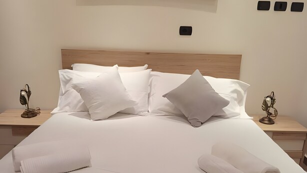 Una cama bien hecha con ropa blanca, dos mesitas de noche con lámparas, y un cuadro abstracto grande sobre la cama.