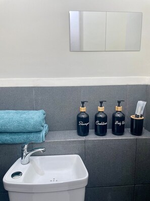Ein Badezimmer mit einem weißen Waschbecken, einem Handtuch und drei schwarzen Flaschen, beschriftet mit 'Shampoo', 'Spülung' und 'Duschgel'.