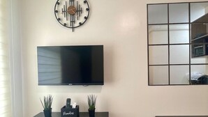 Un salon équipé d’un téléviseur à écran plat, d’une horloge murale, d’une petite table avec une plante et d’une table à manger.