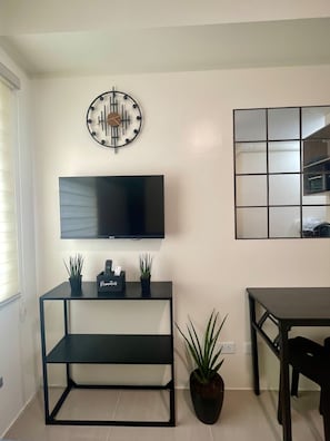 A living room with a flat-screen TV, a wall clock, a small table with a plant, and a dining table. - Studio at Urban Deca Towers Cubao (Quezon City)
