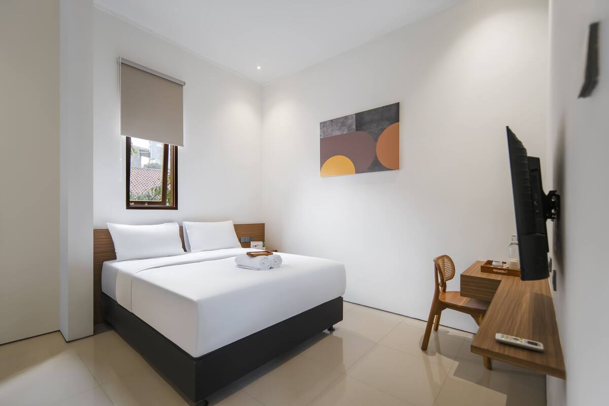 A modern hotel room with a bed, a desk, a chair, a television, and artwork on the wall.
