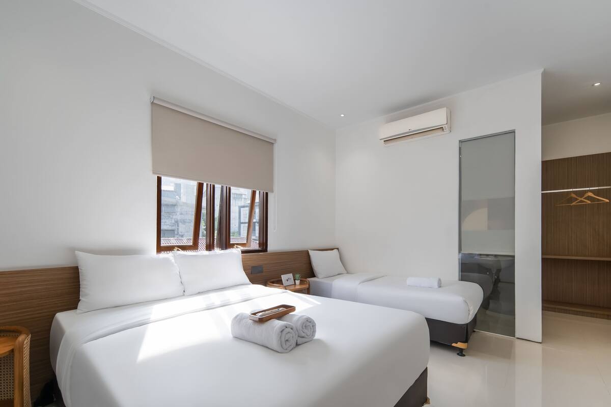 A modern hotel room with two beds, a small table with a card and a plant, a window with curtains, an air conditioning unit, and a wardrobe.