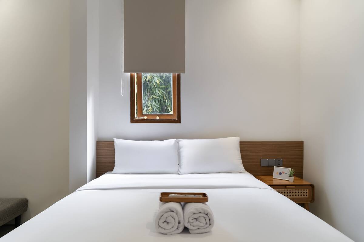A neatly made bed with white linens, a small bedside table with a plant, and a window with a view of greenery.