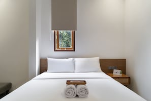 Una cama bien hecha con sábanas blancas, una mesita de noche con una planta, y una ventana con vista a zonas verdes.