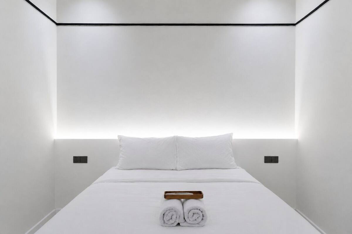 A modern hotel room with a minimalist design, featuring a neatly made bed with white linens and rolled towels, a wooden tray, and recessed lighting.