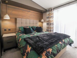 Un dormitorio moderno con cabecera de madera, cama con ropa de cama estampada, mesitas de noche y una ventana con cortinas estampadas.