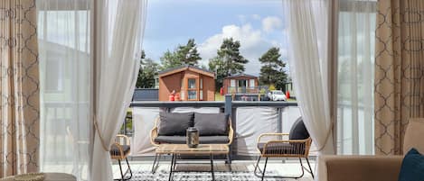 Un área de estar techada al aire libre con una mesa, sillas y un sofá, con vista a edificios al fondo.