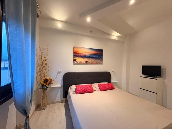 A hotel room with a bed, a TV, a painting of a sunset, and a vase with flowers. - Lake Retreat (Brusimpiano)