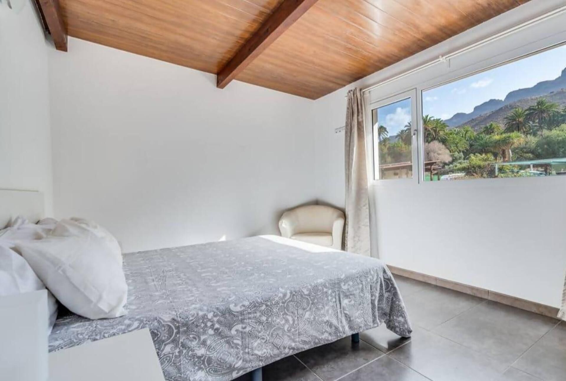 Een slaapkamer met een bed, een stoel, een raam met uitzicht op bergen, en een houten plafond.
