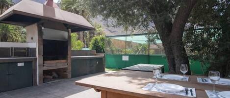 Un patio al aire libre con una parrilla, una mesa lista para comer y vistas a árboles y montañas.